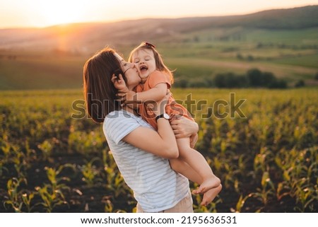 Young mother kissing her baby daughter in the green cornfield on summer day at sunset. Motherhood child care concept. Summer relaxation concept. Summer nature