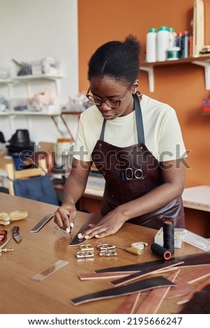 Vertical portrait of black young woman cutting leather and creating handmade belt in workshop