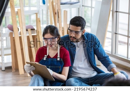 Couple  is checking and inspecting products. Woman hold clip board write plan list in ware house. Young woman and man sit down and hold clipboard work indoors in store.