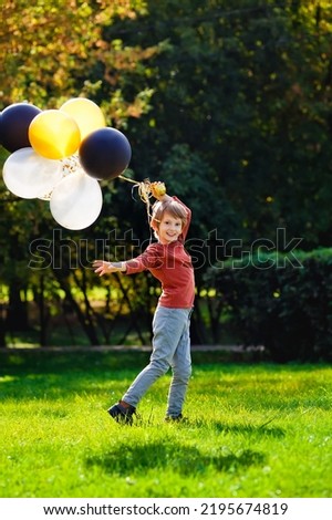 A boy of primary school age runs and jumps with balloons in an autumn sunny park. A red-haired boy with a bunch of multicolored gold, black, and silver balls.