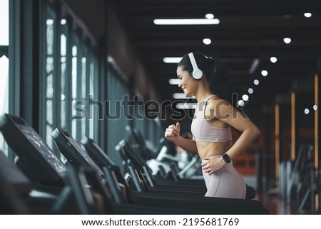 Side view of beautiful young asian woman running on treadmill and listening to music via headphone during sports training in a gym. Royalty-Free Stock Photo #2195681769