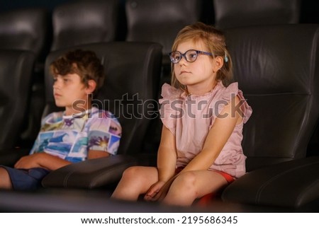 Little preschool girl with glasses and kid boy watching cartoon movie in cinema and eating popcorn. Happy excited children, siblings, brother and sister. Family activity with children.