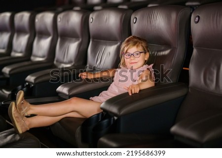 Little preschool girl with glasses watching cartoon movie in cinema and eating popcorn. Happy excited child. Activity with children.