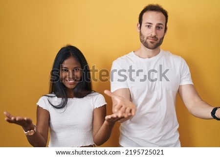 Interracial couple standing over yellow background smiling cheerful offering hands giving assistance and acceptance. 
