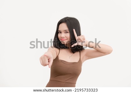 An asian woman wearing a brown tank top teasing you and showing a Loser hand sign crossed isolated on a white background