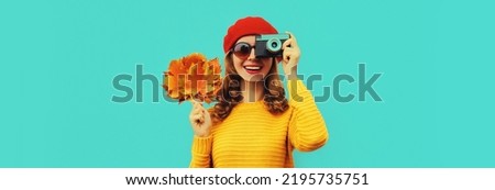 Autumn portrait of happy smiling young woman with film camera and yellow maple leaves wearing sweater, french beret on blue background