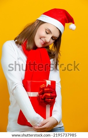 Shy Positive Caucasian Woman in Santa Hat and White Shirt Holding Wrapped Red Gift Box in Front While Looking Downwards Over Yellow Background. Vertical Image Composition