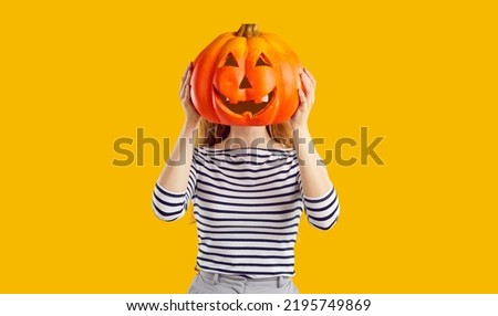 Studio portrait of a woman with a pumpkin head. Funny young girl standing isolated on a yellow background, holding a carved orange pumpkin and hiding her face behind it. Halloween concept Royalty-Free Stock Photo #2195749869