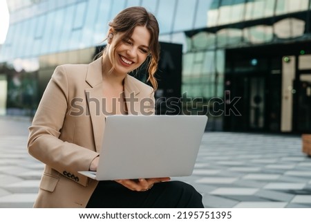 Happy young female blonde female student typing on laptop, talking on video call while sitting outdoors in city, business woman or female freelancer, working outdoors, cityscape in the background.