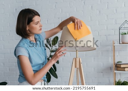 young brunette woman cleaning lamp with rag in living room Royalty-Free Stock Photo #2195755691