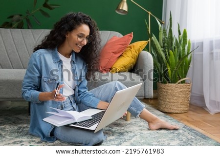 Young beautiful Hispanic female student studying remotely online at home, taking lecture notes sitting on floor in living room with green wall, woman taking courses studying online