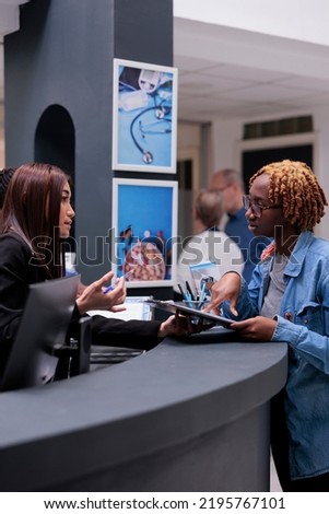 Female receptionist helping woman at reception desk, signing medical report papers to receive insurance support before checkup appointment. Patient filling in registration form to do examination.