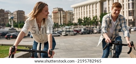 Girl looking at boyfriend while they riding bicycles on city square. Modern urban healthy lifestyle. Entertainment, leisure and hobby. Cycling. Young smiling caucasian couple enjoying time together