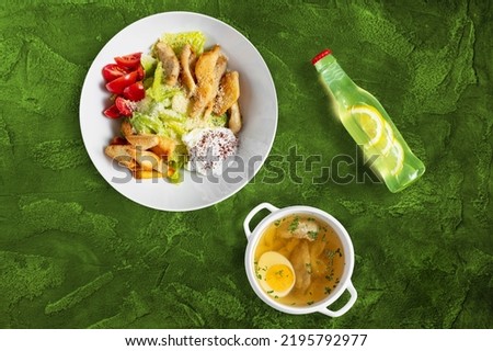 Caesar salad with chicken and chicken broth. Caesar salad with breaded fillet and chicken broth with meatballs. Food lies in light ceramic dishes. Nearby lies lemonade with lemon slice