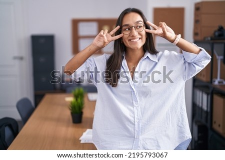 Young hispanic woman at the office doing peace symbol with fingers over face, smiling cheerful showing victory 