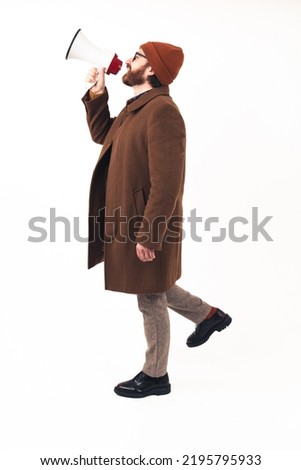 Man in his 30s using megaphone to yell - dressed in a camel coat and stylish boots - profile shot on white background - isolated. High quality photo