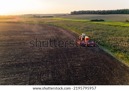 Tractor with sowing machine driving through brown field with greenery on the side and in the fading background. Morning landscape. Horizontal shot. High quality photo