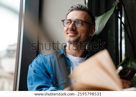 Portrait of adult handsome smiling man in glasses looking out the window while sitting with book in cafe Royalty-Free Stock Photo #2195798531