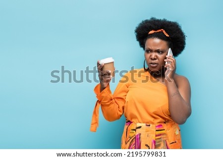 worried african american body positive woman holding paper cup and and talking on smartphone isolated on blue