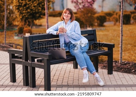 Profile photo of cute lovely young lady sit bench make video call wave hello, greet teacher remote study work online lessons bamboo cup coffee wear white t-shirt jeans park outdoors