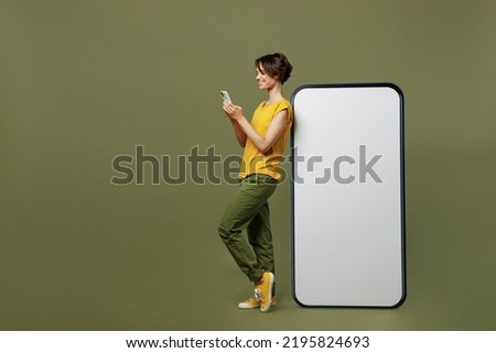 Full body young happy woman she 20s wear yellow t-shirt near big huge blank screen mobile cell phone with workspace copy space mockup area use smartphone isolated on plain olive green khaki background