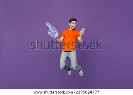 Full body young fan man he wear orange t-shirt cheer up support football sport team hold mobile cell phone 1 fan foam glove finger up watch tv live stream jump isolated on plain dark purple background