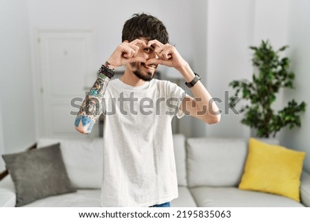 Hispanic man with beard at the living room at home doing heart shape with hand and fingers smiling looking through sign 