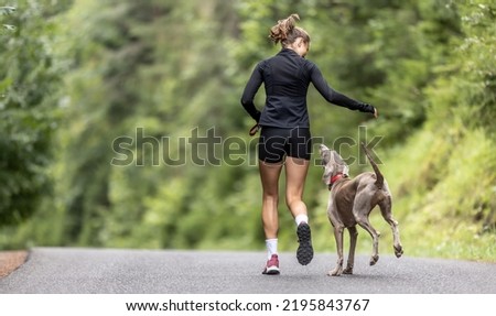 Picture from behind of a girl running in the nature with her dog jumping on her side.