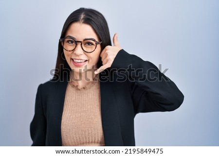 Young brunette woman standing over blue background smiling doing phone gesture with hand and fingers like talking on the telephone. communicating concepts. 