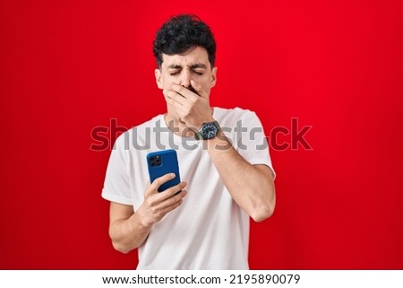 Hispanic man using smartphone over red background bored yawning tired covering mouth with hand. restless and sleepiness. 