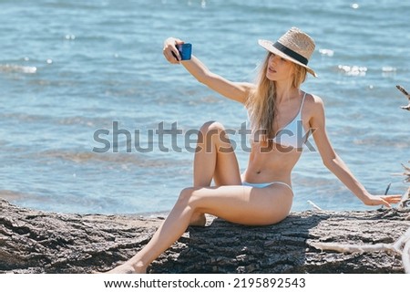 A beautiful long legged blonde girl in a white bikini with hat taking a selfie in front of the sea sitting on a tree trunk