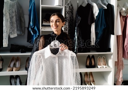 Happy stylish female looking away and smiling while standing in dressing room near closet with many shelves and dressing up at home Royalty-Free Stock Photo #2195893249