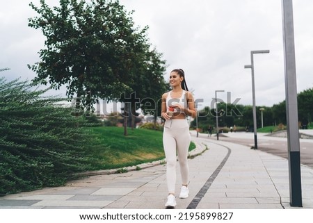 Full length of happy female athlete in sportswear smiling and looking away while walking on paved sidewalk with bottle in hands on street Royalty-Free Stock Photo #2195899827