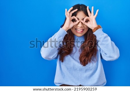 Hispanic young woman standing over blue background doing ok gesture like binoculars sticking tongue out, eyes looking through fingers. crazy expression. 