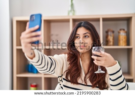 Hispanic young business woman taking a selfie picture drinking a glass of wine relaxed with serious expression on face. simple and natural looking at the camera. 