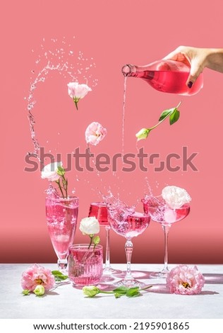 Different elegant champagne glasses with sparkling wine, women hand is pouring rose wine from bottle to coupe glass. Splash, splatter, fly flowers with water drops on the pink background Royalty-Free Stock Photo #2195901865