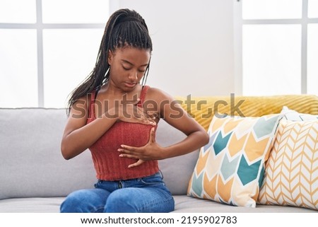 African american woman examining breast with hand sitting on sofa at home Royalty-Free Stock Photo #2195902783