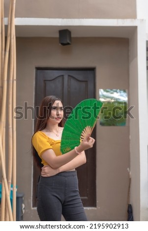 A young asian woman pulls out her big green fan on a humid tropical summer afternoon.