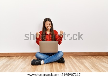 Young brunette woman sitting on the floor at empty room with laptop very happy and excited doing winner gesture with arms raised, smiling and screaming for success. celebration concept. 