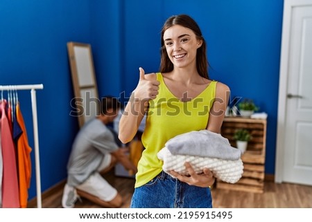 Young brunette woman holding folded laundry smiling happy and positive, thumb up doing excellent and approval sign 
