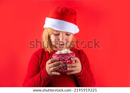 Funny caucasian girl with cup of Christmas cocoa. Happy cute european blonde girl in red sweater and Santa hat hold and drink cup of hot chocolate with marshmallow, on red background