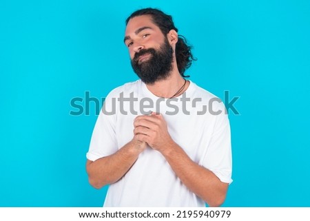 Charming serious Caucasian man with beard wearing white t-shirt over blue background  keeps hands near face smiles tenderly at camera