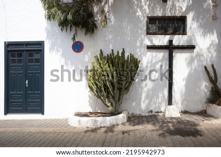 Romantic street with cactus plants and a wooden cross. White wall of the building with a dark blue entrance door. Traffic sign no standing. Teguise, Lanzarote, Canary islands, Spain.
