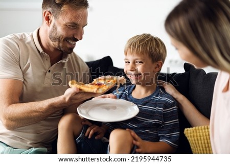 Happy mid adult father feeding his son with pizza at home and little boy  is looking at camera