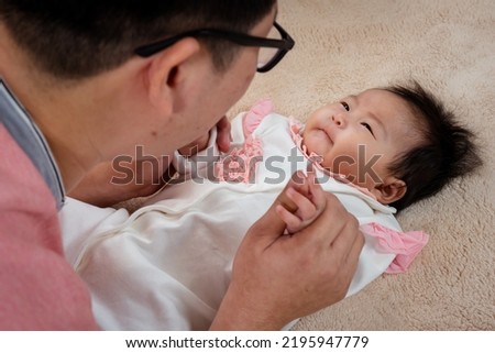 An Asian father in glasses is teasing and playing with his 3 month old newborn daughter in bed in the bedroom with the love. A man who is a single father is taking care of his daughter.