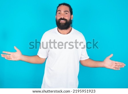 So what? Portrait of arrogant Caucasian man with beard wearing white t-shirt over blue background shrugging hands sideways smiling gasping indifferent, telling something obvious. Royalty-Free Stock Photo #2195951923