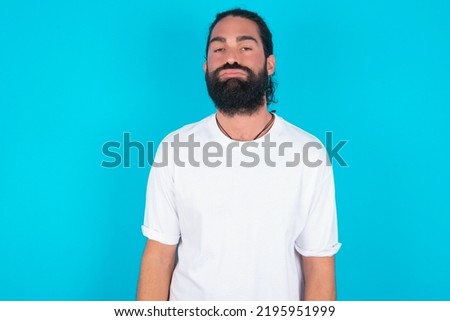 Gloomy, bored Caucasian man with beard wearing white t-shirt over blue background frowns face looking up, being upset with so much talking hands down, feels tired and wants to leave.