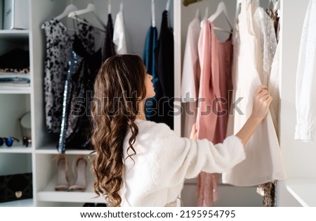 Side view of pensive woman with long dark hair in white sweater standing near closet and choosing clothes on hangers in light wardrobe with various garments Royalty-Free Stock Photo #2195954795