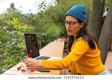 Young student woman with blue hat and yellow sweater writing in a notebook next to her laptop sitting in a park outside