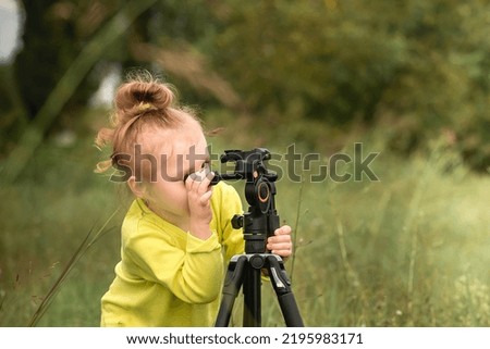 little inquisitive girl with blond wavy hair plays with a tripod for photos in the park in the warm season